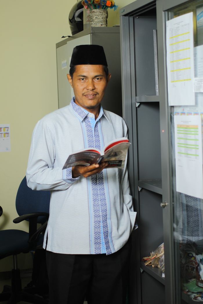 A male teacher in traditional attire holding a book in an office setting.