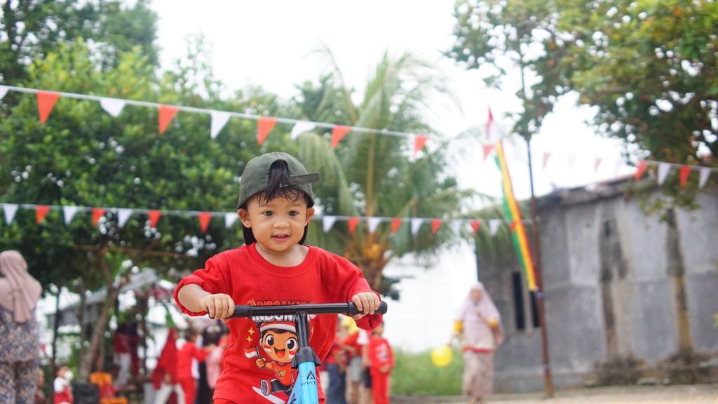 A child riding a scooter during an outdoor festival in Indonesia, with festive decorations and people in the background.