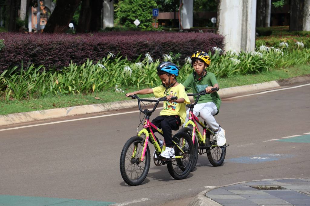 Two children wearing helmets ride bicycles in a sunny Jakarta park.