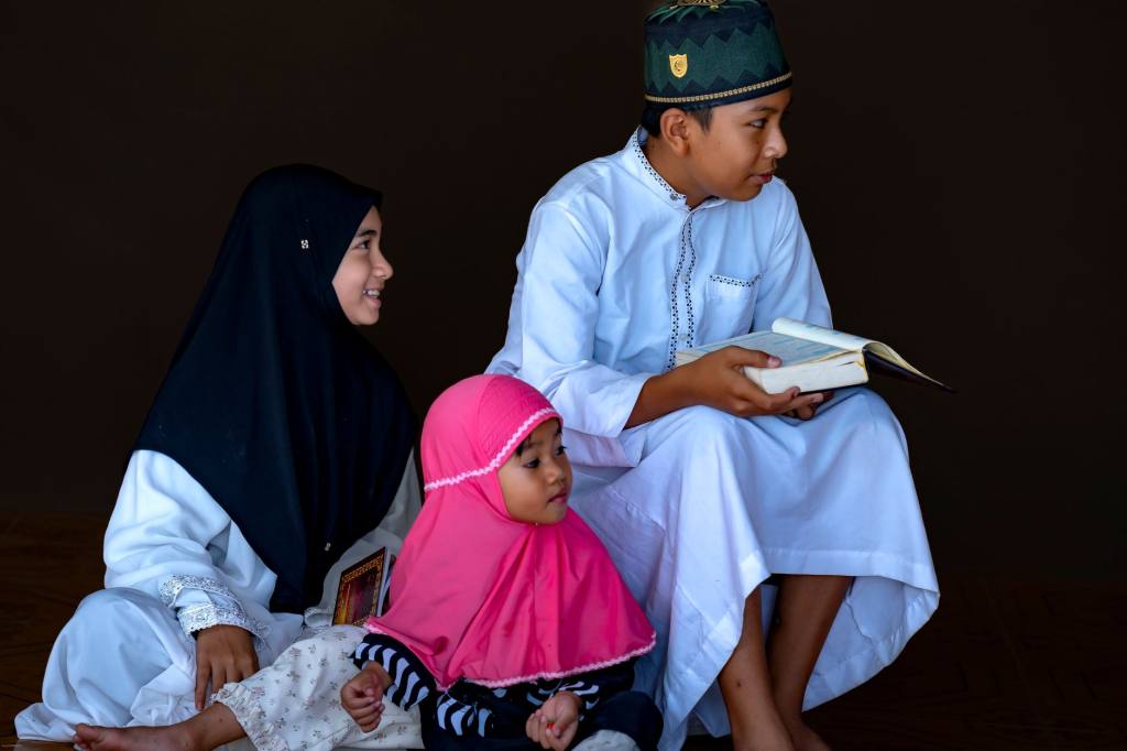 Three children reading the Quran together, wearing traditional outfits.