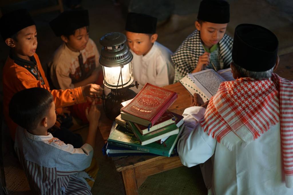 Group of boys studying religious texts around a lamp, guided by an adult indoors.